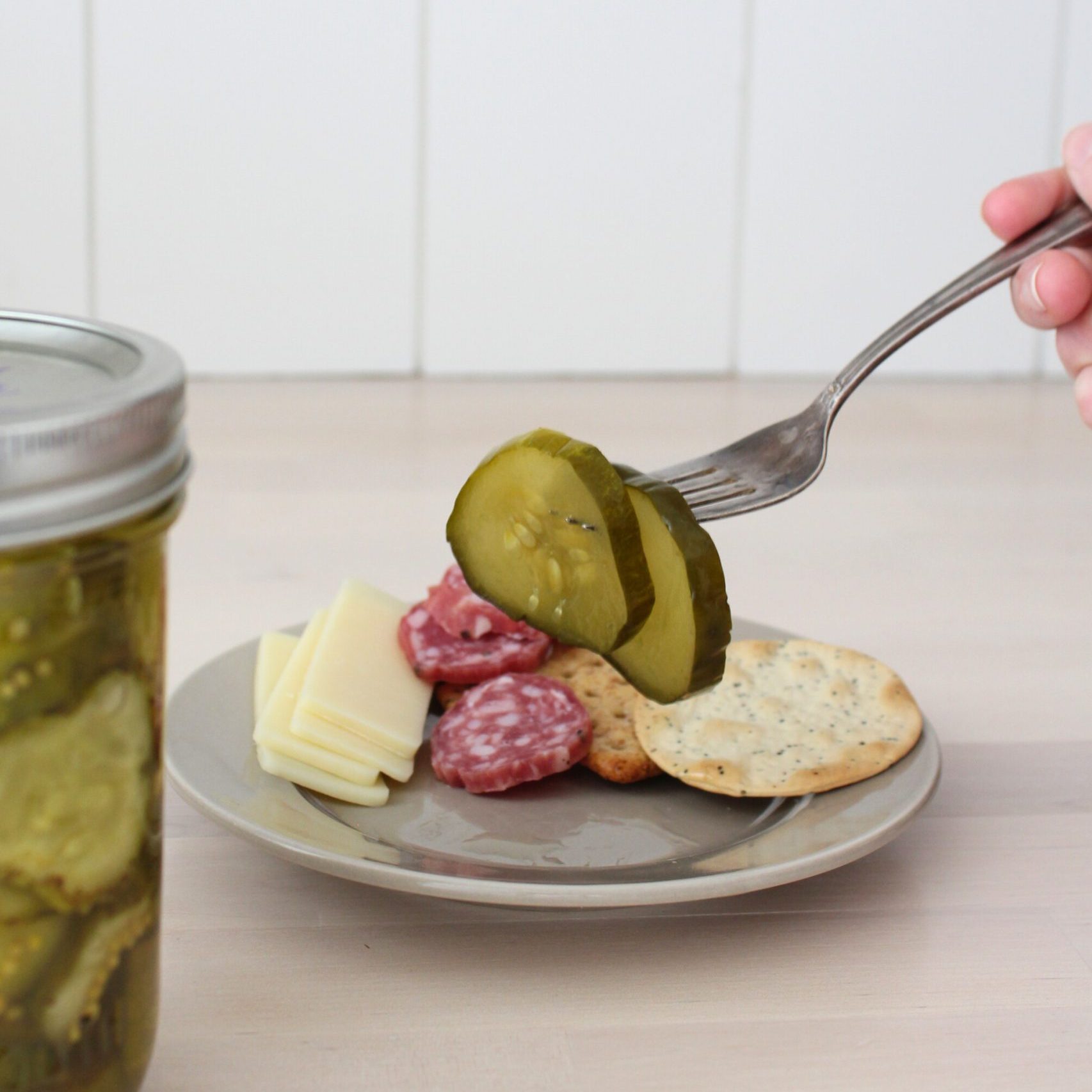 Fork holding up two bread and butter pickles with charcuterie board