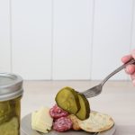 Fork holding up two bread and butter pickles with charcuterie board