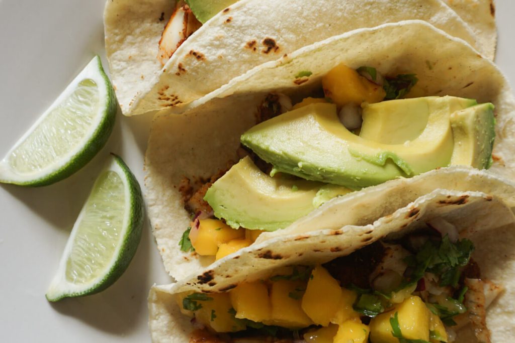 Close up of oven baked blackened fish tacos in corn tortillas topped with sliced avocado and mango salsa