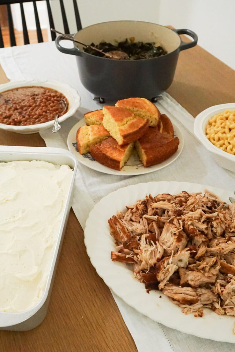 southern barbecue dinner laid out on table with pulled pork, collard greens, Mac and cheese, baked beans, cornbread and banana pudding